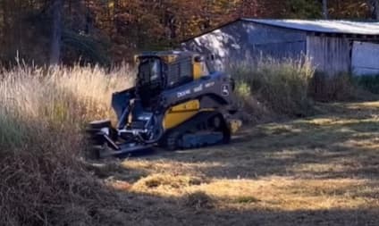 John Deere compact track loader with a brush cutter mowing tall grass beside a barn